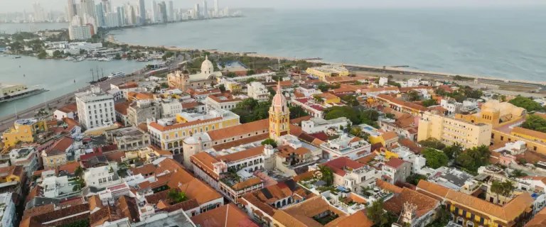 Aerial view of Cartagena’s colonial rooftops and church towers beside the Caribbean coast, with the modern skyline in the distance.