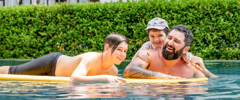 Father and children laughing together on a colorful pool float in a sunny tropical swimming pool.